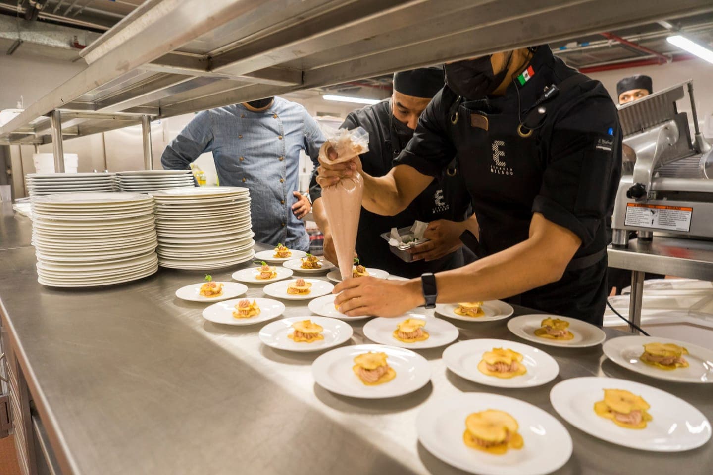 A team of chefs plating a row of dishes on a busy kitchen pass